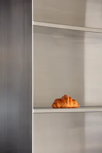 A single croissant sits on a metallic shelf, with a minimalist aesthetic.