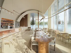Modern restaurant interior with long dining tables, white marble bar, large windows, and soft lighting.