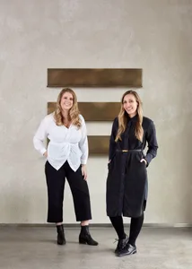 Two women wearing white and black outfits standing against a white wall with gold rectangular panels.