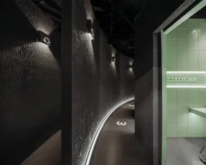 A dark, textured concrete wall with embedded lighting fixtures and a curved corridor leading towards a green-tiled room.