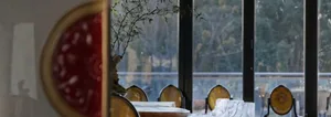 A modern restaurant interior featuring glass windows with a view of an outdoor green area and wooden tables.
