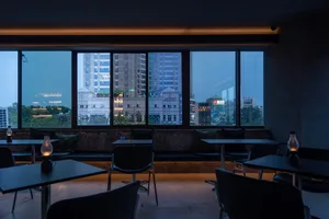 A modern bar with glass windows overlooking a cityscape, featuring concrete furniture and ceiling lights.