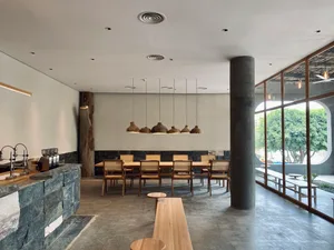 Interior of a minimalist cafe featuring concrete flooring and walls, wooden furniture, and large windows with a view of green