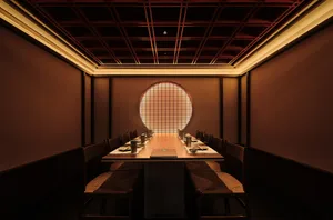 A modern restaurant interior with a long wooden table and chairs, illuminated by warm light from a circular window.