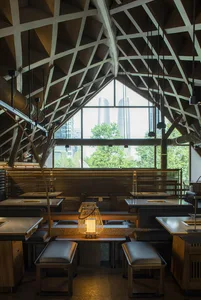 Industrial-style restaurant with wooden tables, metal railings, and glass windows overlooking a cityscape.