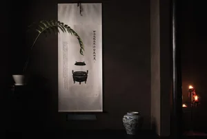 Decorative paper scroll hanging on a dark wall with a plant, vase, and candle holder.