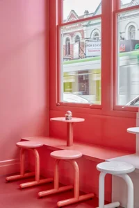 A pink, minimalist restaurant interior with a pink table and stools, and a window overlooking buildings.
