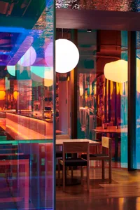 Modern restaurant interior with glass walls, illuminated tables and chairs, and colorful lighting effect.