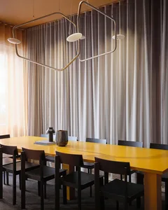 A modern dining room with a yellow wooden table surrounded by black chairs against a backdrop of sheer curtains.