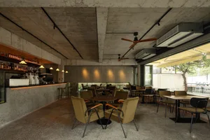 A modern industrial restaurant interior with concrete walls, wooden tables, and chairs, featuring a bar area with hanging lig