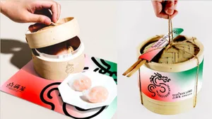 A hand holding a bamboo steamer basket with a tag labeled 'DIAN·MAN LONG' and chopsticks beside it, next to two dumplings on