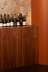 Nine wine bottles lined up on a wooden shelf against a textured wall.