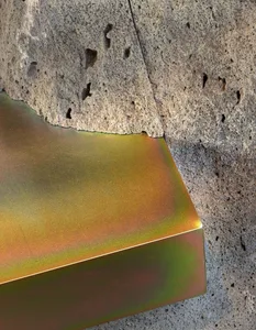 A metallic surface with iridescent hues rests against a concrete wall with holes and cracks.