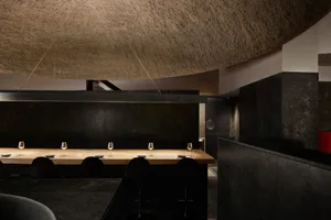 A minimalist restaurant interior with a long wooden table topped with wine glasses and plates, surrounded by black chairs and