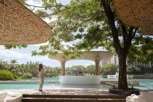 A modern poolside bar with bamboo architecture and a DJ playing music under a large tree.