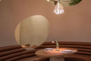 A modern restaurant interior with a curved leather bench seat and a table featuring a vase with a pink flower and cutlery.