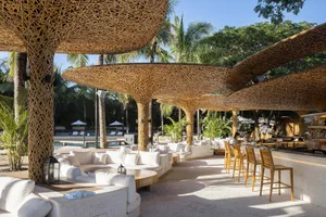 An outdoor lounge area with bamboo ceiling structures and white seating areas surrounded by natural greenery under a clear bl