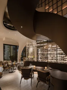 A modern restaurant interior with dark wood tables, glass wine shelves, and natural light through large windows.