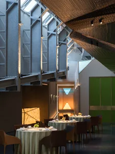 A minimalist restaurant interior features metallic structures, glass walls, and wooden tables and chairs.
