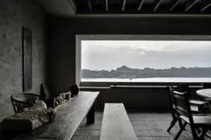 Modern minimalist living room with concrete walls and wooden furniture, overlooking a calm lake and distant hills.