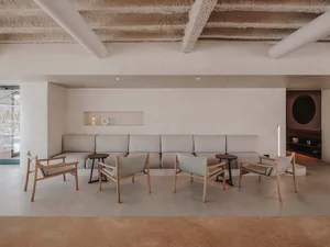 A minimalist living room featuring concrete flooring and a large sofa with wooden armchairs and side tables.