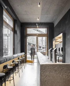 A modern, industrial-style bar with a concrete counter and seating area, featuring a minimalist design and a glass door.
