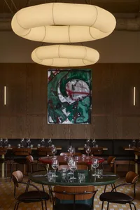 Modern restaurant interior with wood and metal tables and chairs, glass lighting fixtures, and abstract painting on the wall.
