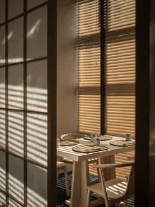 Restaurant interior with wooden chairs and blinds casting stripes on the wall.