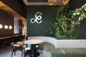 A modern industrial coffee shop interior with green wall, curved seating, and potted plants.