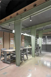 An industrial-style restaurant interior with metallic columns, corkboard walls, and wooden tables.