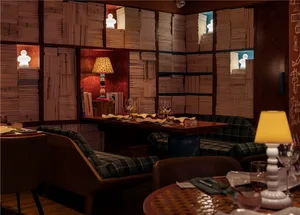 A modern restaurant bar library with wooden walls, paper books arranged on shelves, and a white lamp on a wooden table.