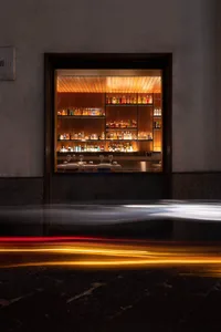An illuminated restaurant bar with shelves filled with bottles and glasses, set against a dark exterior wall with blurred lig
