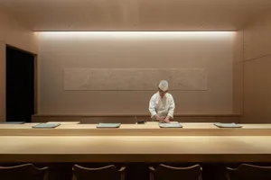 A chef in a traditional chef's hat prepares food behind a long wooden counter in a minimalist restaurant interior.