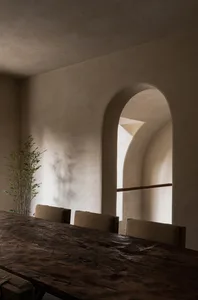 A wooden dining table with chairs in a restaurant setting with arched windows, concrete walls, and a small potted plant.