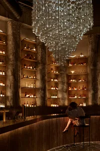 A dark and moody interior bar with concrete walls and shelves filled with bottles of alcohol. A woman sits in the foreground 
