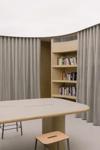 A modern study room with a wooden table and shelves filled with books, enclosed by fabric curtains and a wooden cabinet.