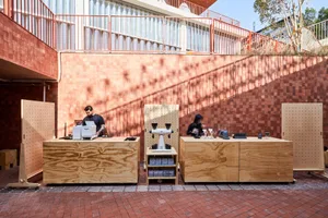 Two coffee shop employees working at wooden stations with coffee machines and equipment on a brick patio with a red wall in t