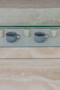 Two gray ceramic mugs and three white ceramic cups arranged on a glass shelf.