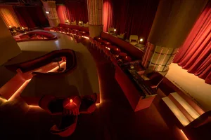 A modern bar with red leather seats and tables, featuring a stage and seating area.