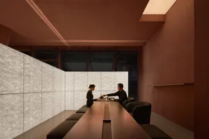 Two individuals sitting at a long wooden table in a modern coffee shop with a concrete wall and wooden flooring.