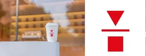 A white paper coffee cup with a red triangle and square logo sits on a white table in a modern coffee shop.