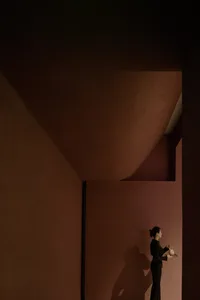 A minimalist interior space featuring exposed concrete walls and a single figure holding a teapot on a tray.