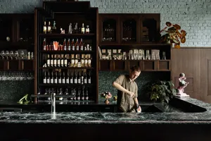 A man working in a restaurant bar with a stone counter, wood shelves, and various bottles and glasses.
