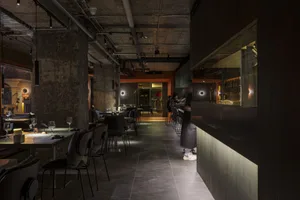 A modern, industrial restaurant interior with concrete walls, metal accents, and glass windows.