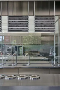A modern industrial restaurant interior featuring a metal and glass bar counter with digital signs.