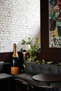 A modern bar with white brick walls, featuring a large champagne bottle, a green plant, and a lamp on a wooden floor.