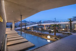 An architectural space featuring a concrete and glass landscape pavilion with a pool, benches, and palm trees at dusk.
