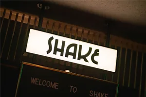 A neon sign with the word 'SHAKE' mounted above a black-framed message board.
