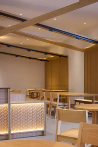 A modern restaurant interior featuring wooden tables and chairs, illuminated by strategically placed lights within the ceilin