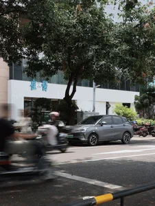 A modern urban scene featuring a grey car parked under a tree, with other vehicles and buildings in the background.